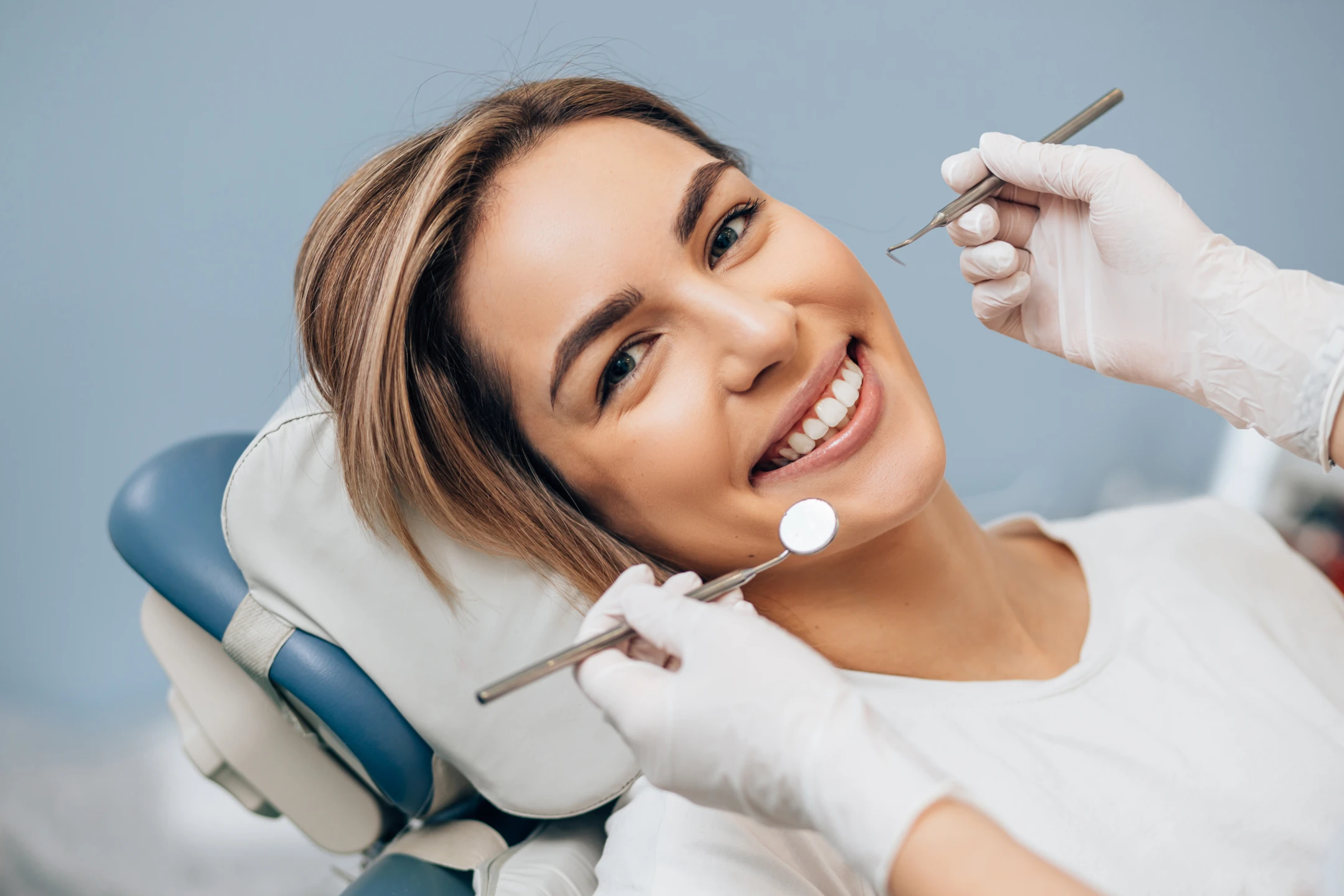 Smiling woman reclines in dental chair during checkup with tools near her mouth.